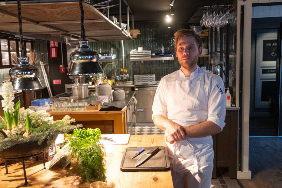 a chef in a kitchen standing next to a chopping board and two knives