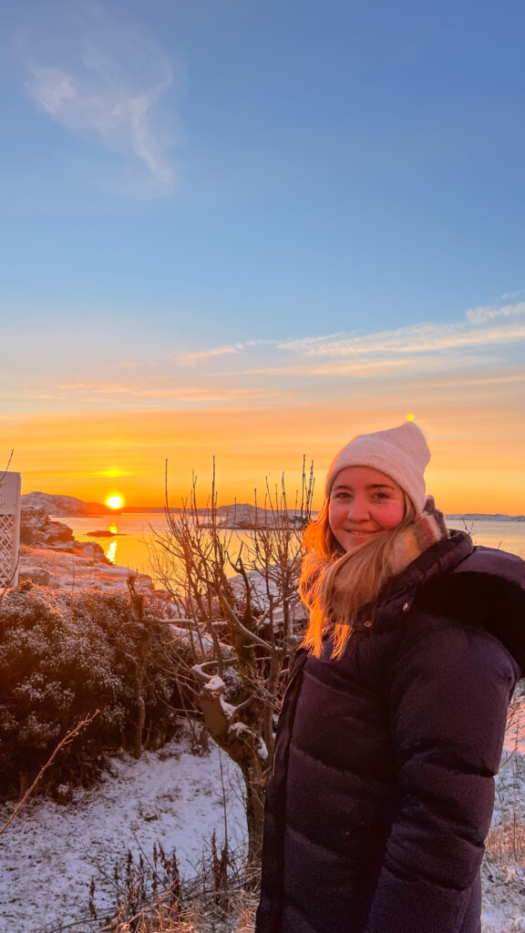 en smilende kvinne med vinterklær på foran soloppgang over havet og snø på bakken